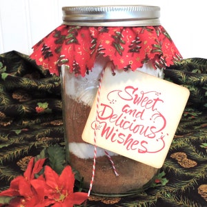 May include: A glass jar filled with ingredients for a baking mix, decorated with a red and green fabric trim and a tag that reads "Sweet and Delicious Wishes".