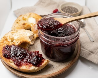 May include: A glass jar filled with dark red berry jam containing visible chia seeds, with a wooden spoon lifting a portion of the jam. Beside the jar, a toasted bagel half is spread with the same jam, and another bagel half shows melted butter.