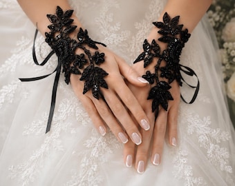 May include: A pair of black lace fingerless gloves featuring intricate floral embroidery, small black sequins, and thin black ribbon ties. The lace design extends from the wrist to the back of the hand, creating a decorative pattern over the skin.