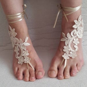 May include: A pair of champagne-colored barefoot sandals featuring intricate floral lace embroidery. Each sandal includes a delicate ribbon tie that wraps around the ankle and a thin loop that secures around the second toe, showcasing a detailed vine pattern.