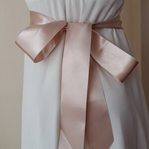 May include: A blush pink satin ribbon tied in a bow around a white dress.