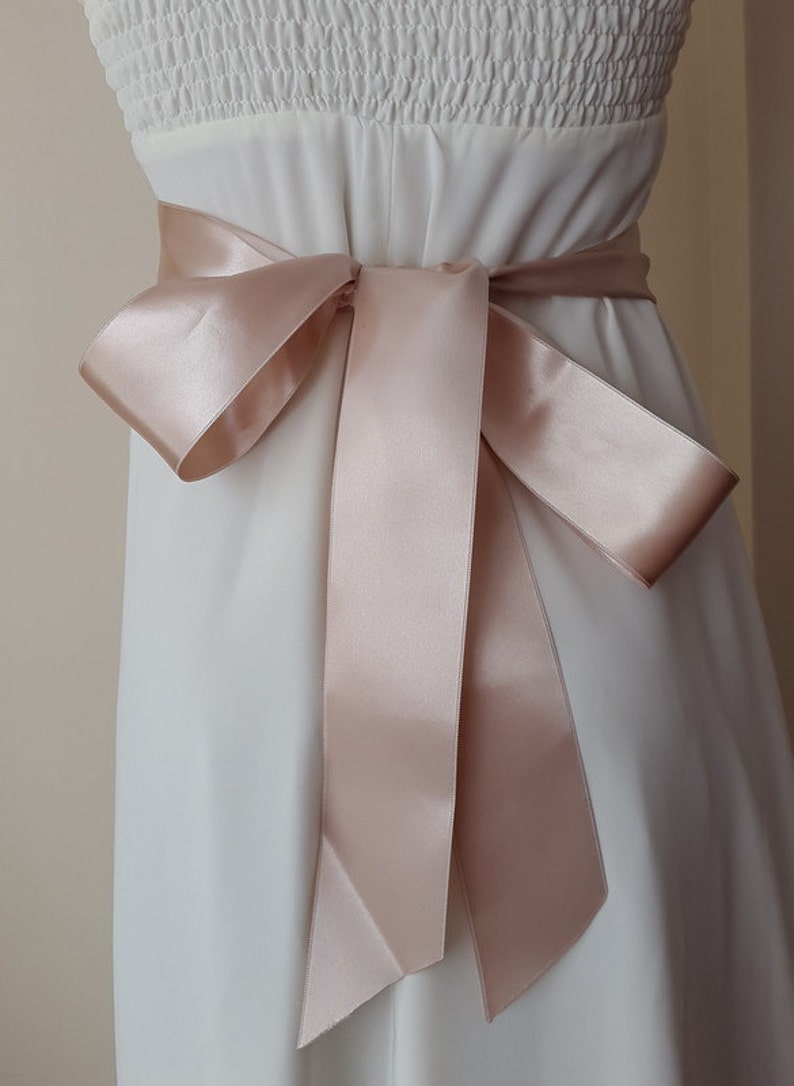 May include: A blush pink satin ribbon tied in a bow around a white dress.