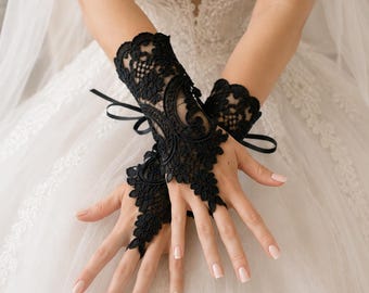 May include: A pair of black lace fingerless gloves featuring intricate floral embroidery and scalloped edges. The gloves are designed to cover the wrist and back of the hand, secured with a delicate black ribbon tie at the wrist and a loop for the middle finger.