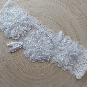 May include: White lace garter with three white floral appliques and rhinestones.