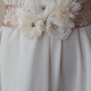 May include: A white satin bridal sash with delicate lace trim and three white fabric flowers. The flowers have pearl and rhinestone embellishments.