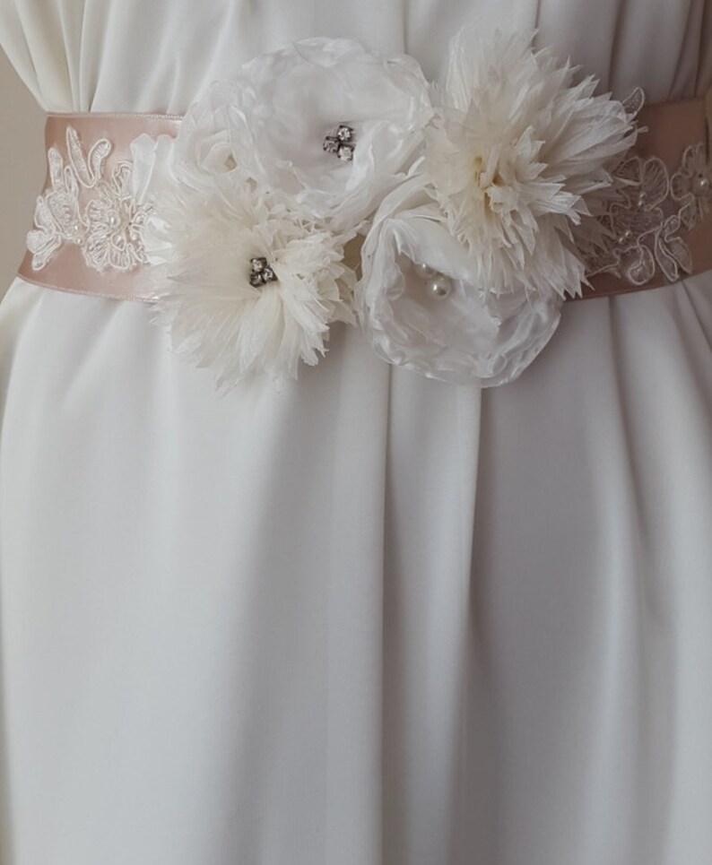 May include: A white satin bridal sash with delicate lace trim and three white fabric flowers. The flowers have pearl and rhinestone embellishments.