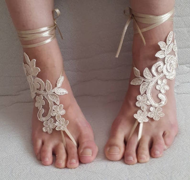 May include: A pair of champagne-colored barefoot sandals featuring intricate floral lace embroidery. Each sandal includes a delicate ribbon tie that wraps around the ankle and a thin loop that secures around the second toe, showcasing a detailed vine pattern.
