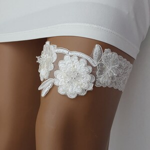 May include: White lace garter with floral embellishments. The garter features two large white flowers with pearl accents and smaller white flowers.