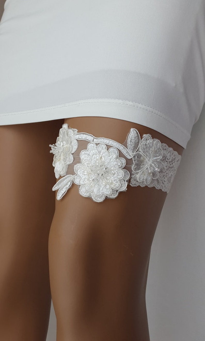 May include: White lace garter with floral embellishments. The garter features two large white flowers with pearl accents and smaller white flowers.