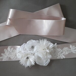 May include: A white satin sash with delicate lace embroidery and a cluster of white fabric flowers. The sash is tied in a bow and has a blush pink satin ribbon underneath.