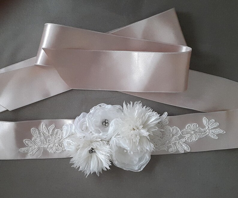 May include: A white satin sash with delicate lace embroidery and a cluster of white fabric flowers. The sash is tied in a bow and has a blush pink satin ribbon underneath.