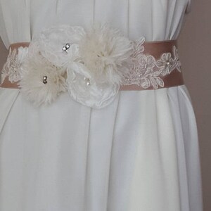 May include: A white satin sash with a delicate lace overlay and a cluster of white fabric flowers. The sash is adorned with small, clear rhinestones.