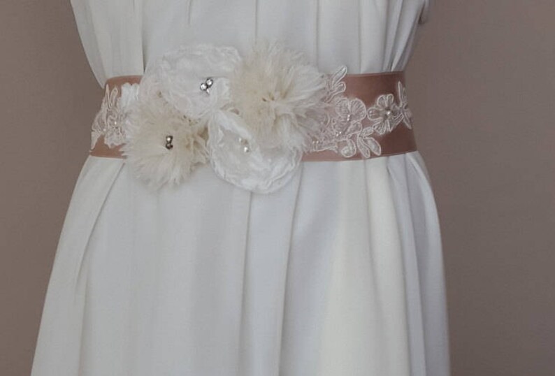 May include: A white satin sash with a delicate lace overlay and a cluster of white fabric flowers. The sash is adorned with small, clear rhinestones.