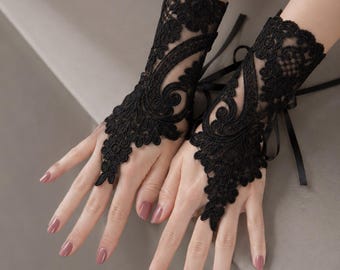 May include: A pair of black fingerless lace gloves featuring intricate floral embroidery and scalloped edges. The gloves extend to the wrist and include a delicate black ribbon tie closure at the back, leaving the fingers and thumb exposed.