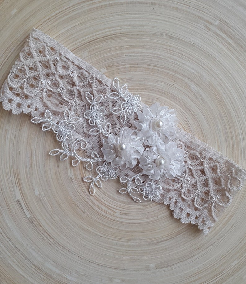 Puede incluir: Una liga de encaje blanco con flores de sat&eacute;n blanco y detalles de perlas.