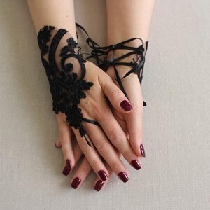 May include: A black lace fingerless glove featuring intricate floral embroidery and swirling patterns. The accessory is designed to cover the back of the hand and includes thin black ribbon ties that wrap around the wrist for a secure fit.