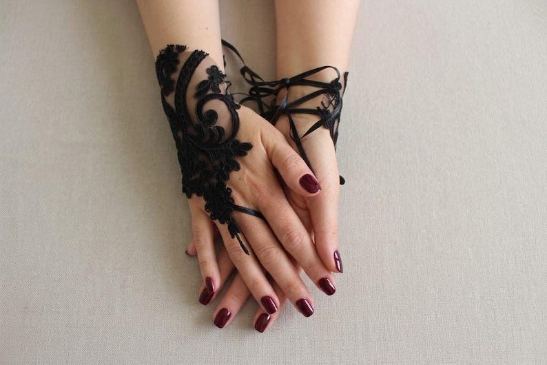 May include: A black lace fingerless glove featuring intricate floral embroidery and swirling patterns. The accessory is designed to cover the back of the hand and includes thin black ribbon ties that wrap around the wrist for a secure fit.