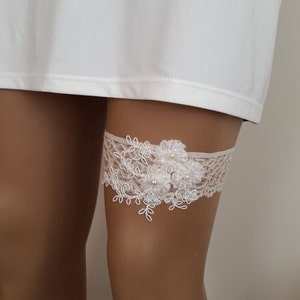 Puede incluir: Jarretera de encaje blanco con un dise&ntilde;o floral y detalles de perlas.