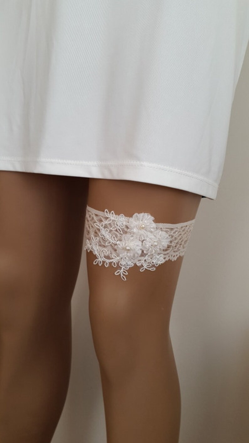 Puede incluir: Jarretera de encaje blanco con un dise&ntilde;o floral y detalles de perlas.