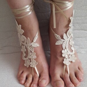May include: A pair of ivory lace barefoot sandals featuring intricate floral embroidery and delicate vine patterns. Each sandal includes a thin satin ribbon that wraps around the ankle and a loop that secures around the second toe, creating a decorative design.