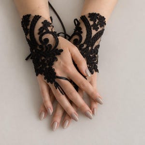 May include: A pair of black fingerless lace gloves featuring intricate floral embroidery and swirling patterns. The gloves are crafted from sheer black mesh fabric and include a delicate ribbon tie detail that secures around the middle finger.