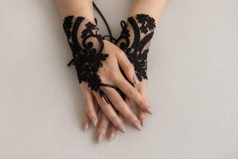 May include: A pair of black fingerless lace gloves featuring intricate floral embroidery and swirling patterns. The gloves are crafted from sheer black mesh fabric and include a delicate ribbon tie detail that secures around the middle finger.