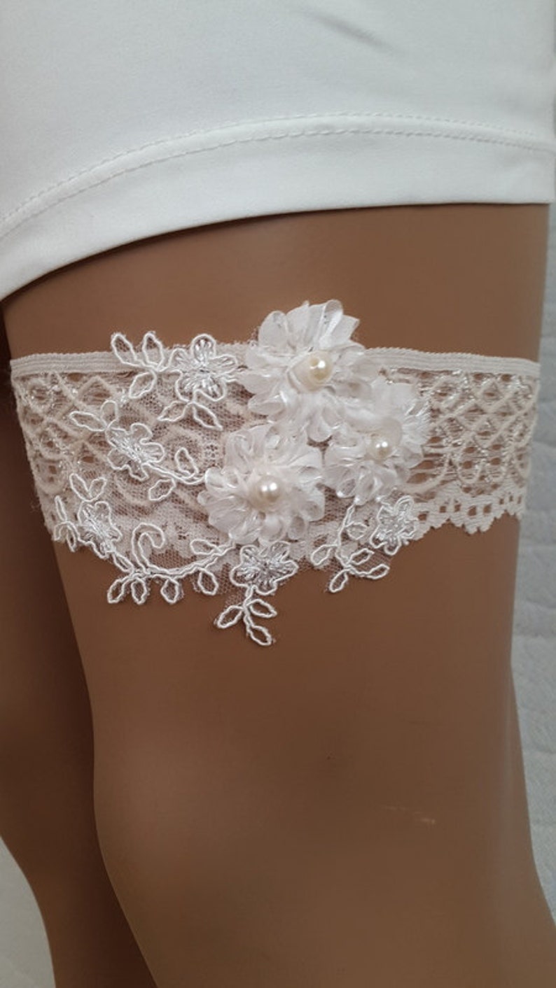 Puede incluir: Jarretera de encaje blanco con tres flores de tela blanca y detalles de perlas.
