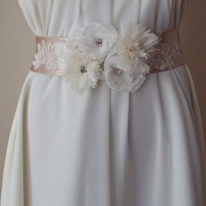 May include: A white satin bridal sash with delicate lace trim and three white fabric flowers adorned with rhinestones.