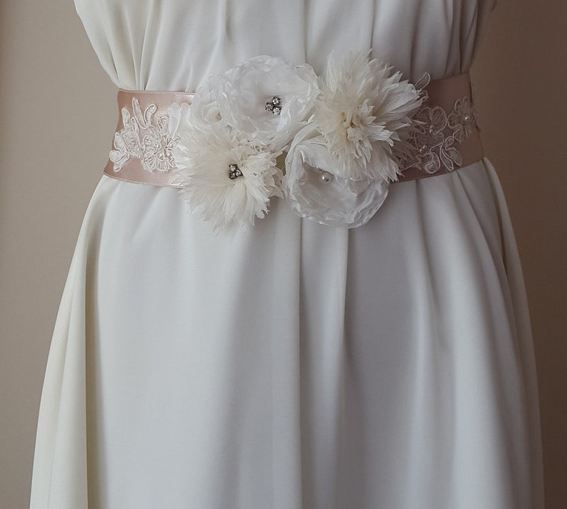 May include: A white satin bridal sash with delicate lace trim and three white fabric flowers adorned with rhinestones.