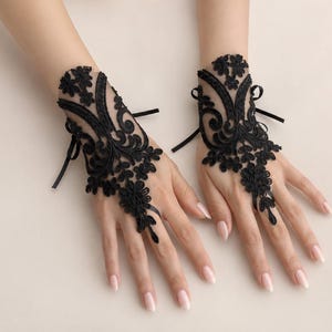 May include: A pair of black lace fingerless gloves featuring intricate floral embroidery and scrollwork patterns. The gloves are designed with a sheer mesh base, a loop for the middle finger, and thin black ribbon ties at the wrist for closure.