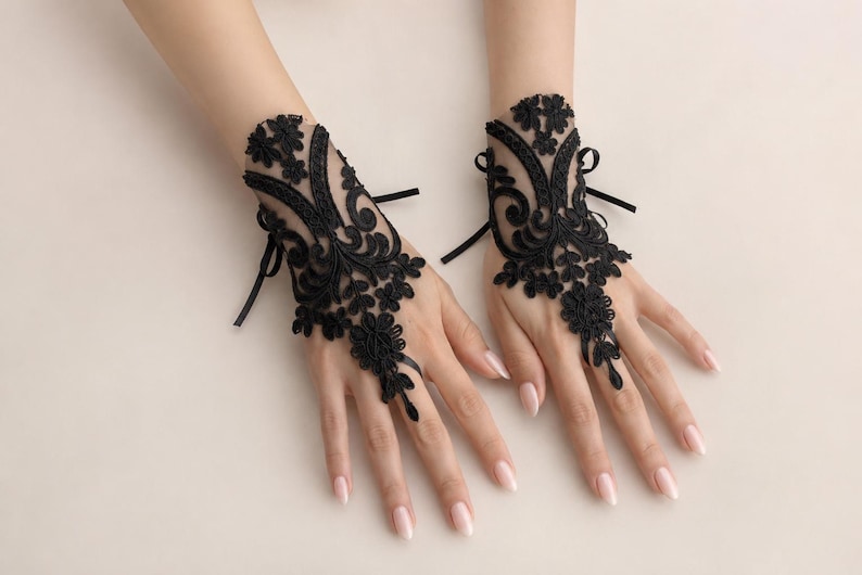 May include: A pair of black lace fingerless gloves featuring intricate floral embroidery and scrollwork patterns. The gloves are designed with a sheer mesh base, a loop for the middle finger, and thin black ribbon ties at the wrist for closure.
