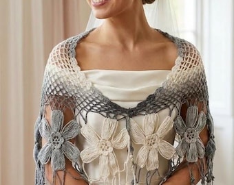 May include: A crochet shawl featuring an ombre colour transition from cream to grey. The garment has an open mesh pattern and is adorned with large, dimensional floral motifs in cream and grey, finished with a fringed edge along the bottom.