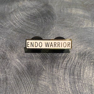 May include: A gold-colored pin with a white background and the text "ENDO WARRIOR" in black lettering.
