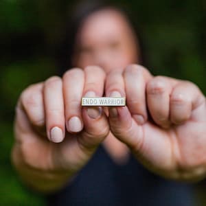 May include: A person's hands holding a gold bar pin with the words "Endo Warrior" written in white.