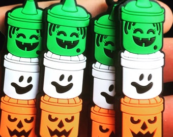 May include: A collection of Halloween-themed keychains. Each keychain features a stacked design with a green witch's hat, a white ghost, and an orange jack-o'-lantern. The keychains have black loops at the top for attachment.