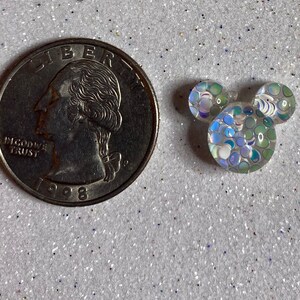 Op de afbeelding: Een kleine, witte, iriserende Mickey Mouse-kopbedel van hars met een blauw en groen glitterpatroon. De bedel is ongeveer zo groot als een Nederlandse euro.