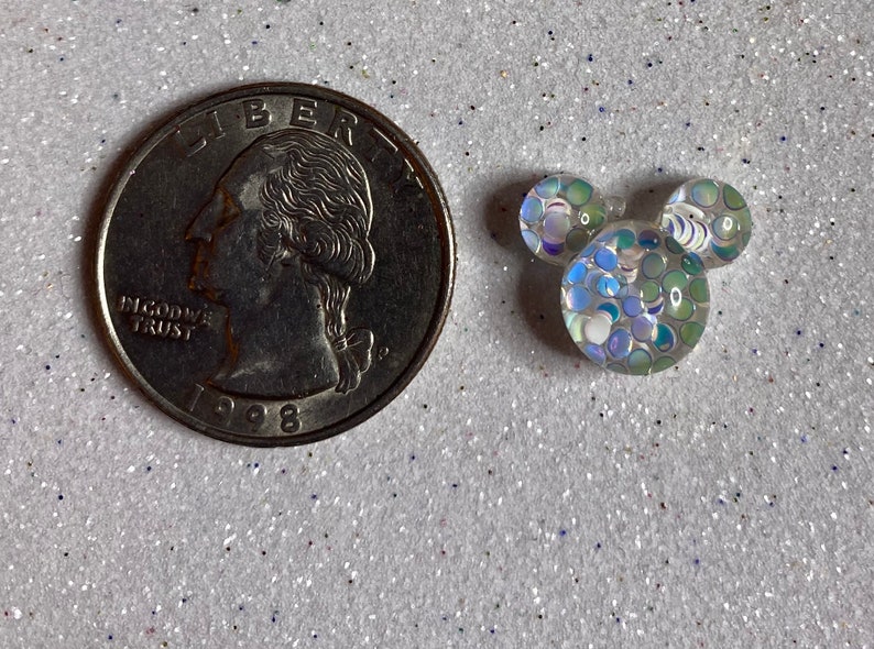 Op de afbeelding: Een kleine, witte, iriserende Mickey Mouse-kopbedel van hars met een blauw en groen glitterpatroon. De bedel is ongeveer zo groot als een Nederlandse euro.