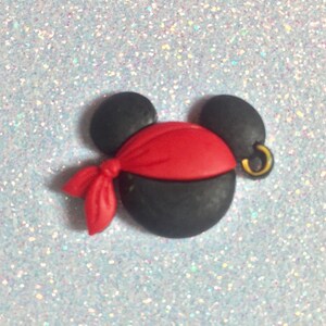 May include: A black and red pirate-themed mouse head with a gold earring. The mouse head has black ears, a black face, and a red bandana. The earring is gold. The background is a light blue glitter.