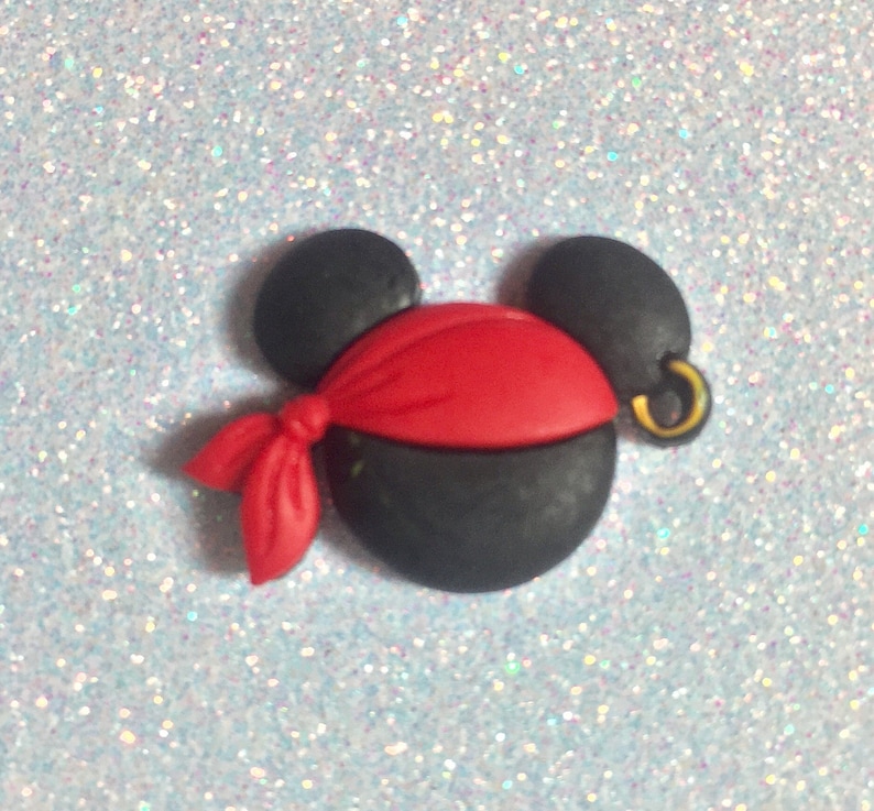 May include: A black and red pirate-themed mouse head with a gold earring. The mouse head has black ears, a black face, and a red bandana. The earring is gold. The background is a light blue glitter.