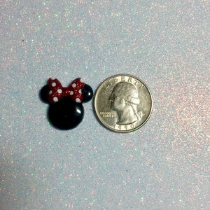 May include: A small black and red Minnie Mouse head with a white polka dot bow. The Minnie Mouse head is next to a US quarter for size comparison.