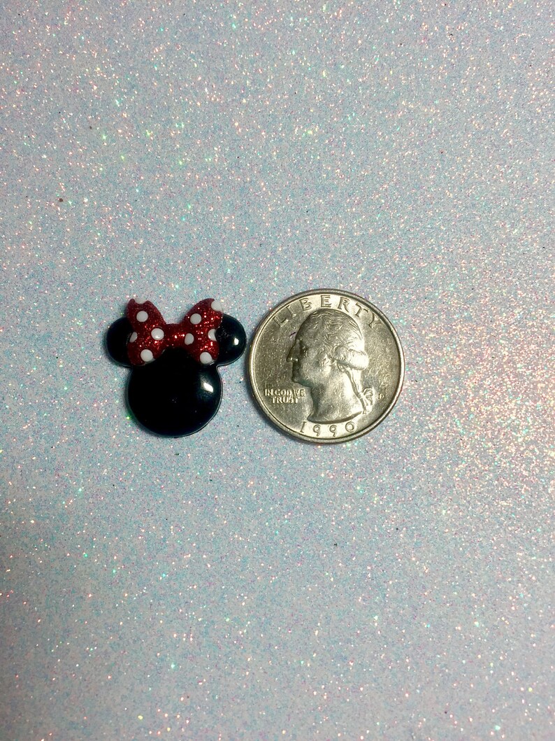 May include: A small black and red Minnie Mouse head with a white polka dot bow. The Minnie Mouse head is next to a US quarter for size comparison.