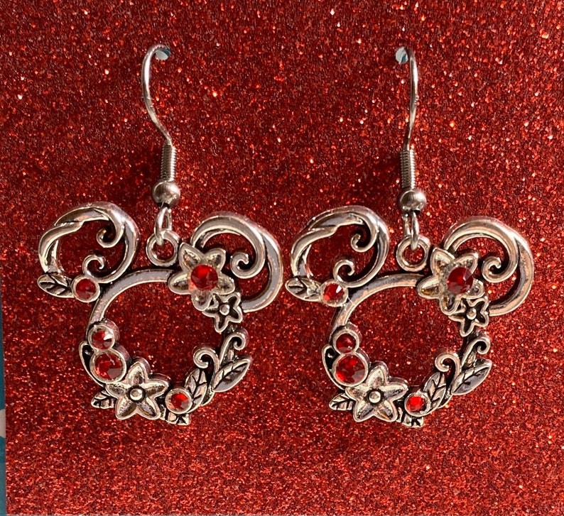 May include: A pair of silver earrings featuring a red rhinestone Mickey Mouse head design. The earrings are made of metal and have a floral design.