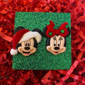 May include: A pair of Christmas-themed Mickey Mouse and Minnie Mouse earrings. Mickey is wearing a red Santa hat and Minnie has a red bow with white polka dots.
