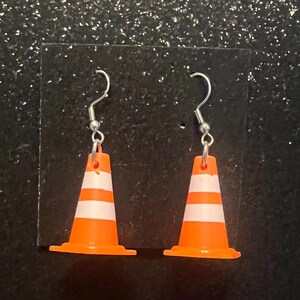 May include: A pair of novelty earrings featuring miniature orange and white traffic cones. The earrings have silver-colored fish hook ear wires. The cones have two white stripes and a wide base.