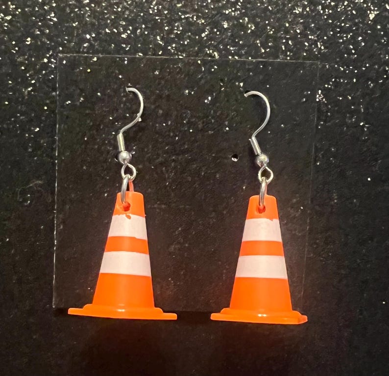 May include: A pair of novelty earrings featuring miniature orange and white traffic cones. The earrings have silver-colored fish hook ear wires. The cones have two white stripes and a wide base.