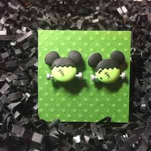 May include: A pair of green and black Frankenstein-themed earrings. The earrings are shaped like Mickey Mouse heads with green faces and black hair. The earrings are on a green background with white polka dots.