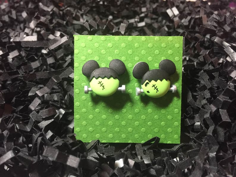 May include: A pair of green and black Frankenstein-themed earrings. The earrings are shaped like Mickey Mouse heads with green faces and black hair. The earrings are on a green background with white polka dots.