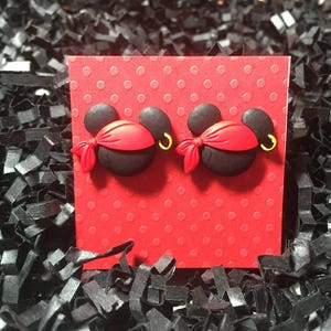 May include: A pair of black and red pirate-themed earrings. Each earring features a black mouse head with a red bandana, a gold hoop earring, and a red bow. The earrings are displayed on a red polka-dotted card.