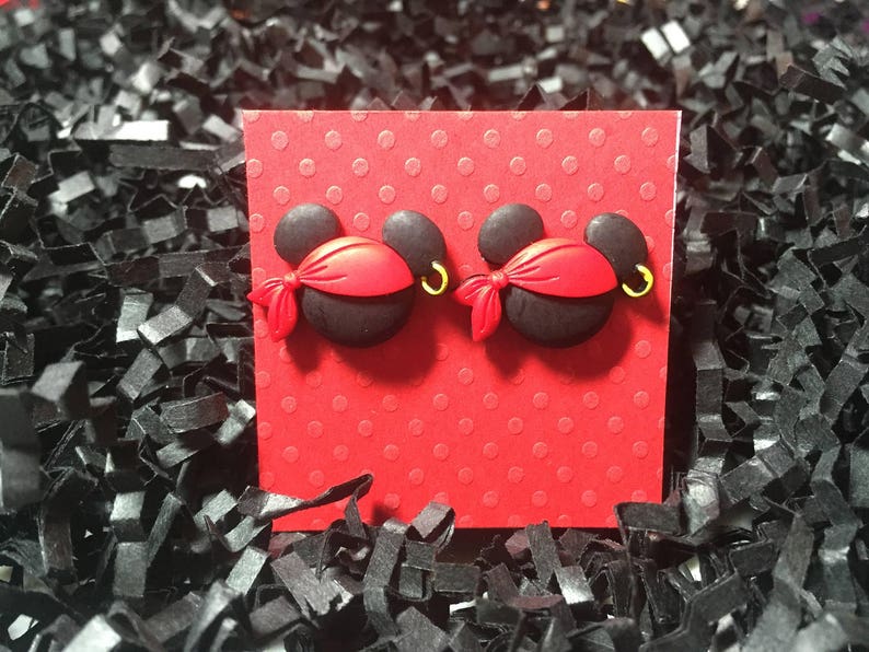 May include: A pair of black and red pirate-themed earrings. Each earring features a black mouse head with a red bandana, a gold hoop earring, and a red bow. The earrings are displayed on a red polka-dotted card.