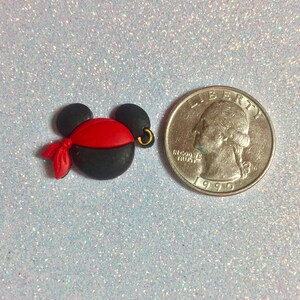 May include: A black and red pirate-themed Mickey Mouse head charm with a gold hoop earring. The charm is next to a U.S. quarter coin for size comparison. The charm has a red bandana and is made of a smooth material.